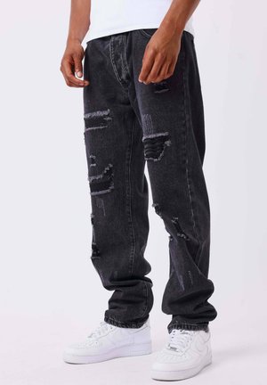 Homme portant un jean noir déchiré ample et des baskets blanches, debout les mains détendues le long du corps devant un fond uni.