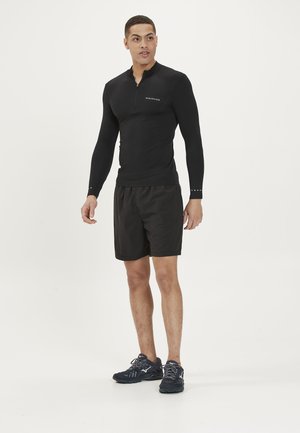 Sort langærmet atletisk top med kvart lynlåskrave, parret med sorte shorts og mørke løbesko. Har diskret branding.