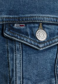 Denim w jasnoniebieskim kolorze, z mosiężnym guzikiem i małym haftowanym logo w kolorach czerwonym, białym i niebieskim na kieszeni.