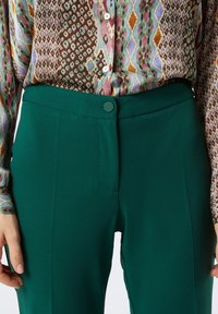 Pantalon vert à texture lisse, coupe ajustée, avec une fermeture à bouton. Associé à une chemise à manches longues colorée et à motifs.