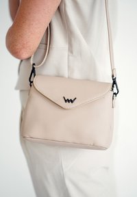 Bežová kabelka přes rameno z umělé kůže s klopou, kontrastním prošíváním a černým logem. Obsahuje nastavitelný popruh a ostré tvary.