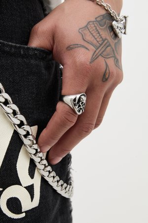 Bague en argent en forme de crâne avec une forme triangulaire, portée sur une main avec un tatouage sombre. Inclut une chaîne en argent attachée à un tissu en denim noir.