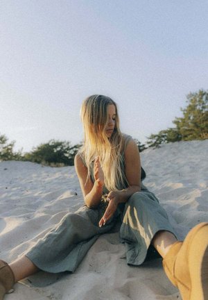 Modelka w jasnooliwkowym kombinezonie siedząca na piaszczystym terenie, delikatnie strzepująca piasek z rąk, z długimi, prostymi włosami i brązowymi butami.