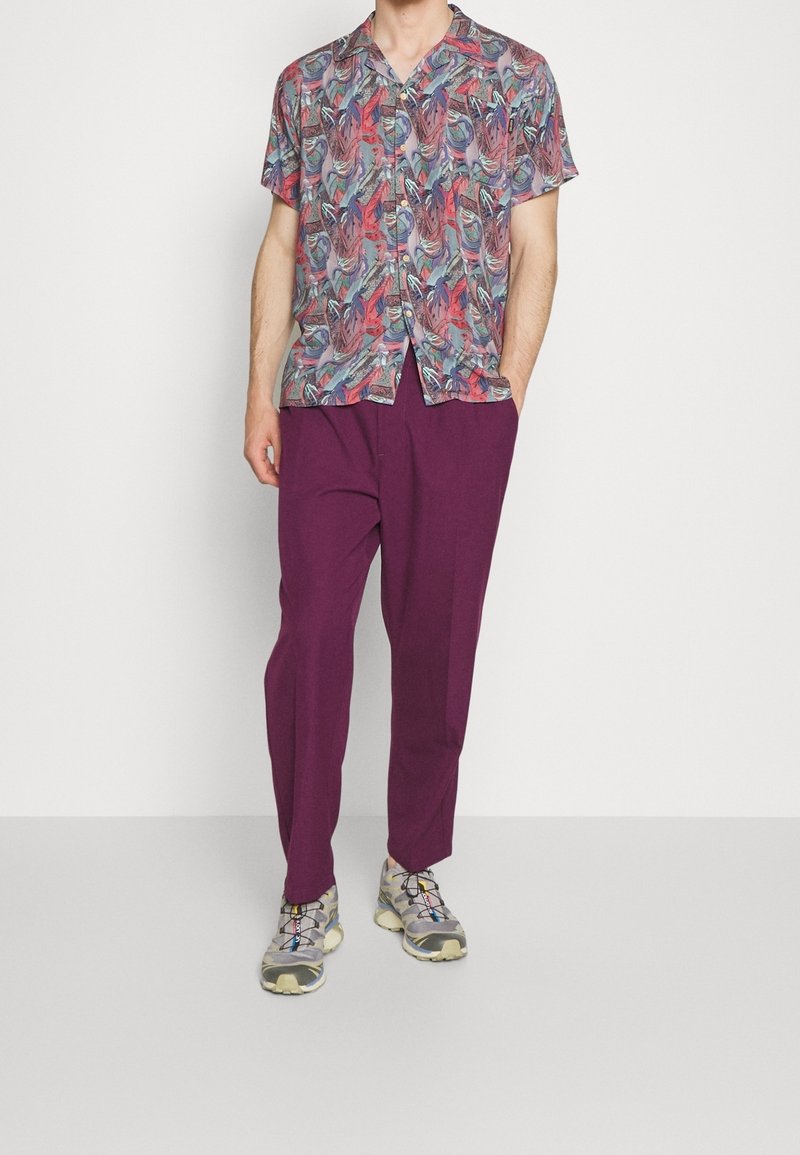 Homme portant une chemise à manches courtes boutonnée avec un motif abstrait rouge et bleu, un pantalon violet foncé et des baskets grises avec des lacets colorés.