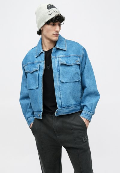 Chaqueta vaquera azul con dos bolsillos frontales, camiseta negra debajo, pantalones grises y un gorro de color claro con un diseño gráfico.