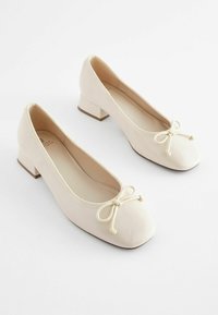 Chaussures de ballet à talons bloc en cuir verni beige avec un bout arrondi et un nœud décoratif sur le devant. Texture lisse et design minimal.