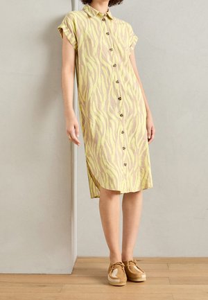 Kobieta ubrana w żółto-beżową sukienkę na guziki o długości za kolano i krótkich rękawach, nosząca mokasyny, stoi na tle jednolitej ściany.