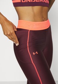 Kobieta nosząca bordowe i pomarańczowe legginsy Under Armour z wysokim stanem, pokazująca boczną kieszeń, w której trzyma rękę.