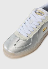 Metāla sudraba kedas ar baltām šņorēm, ar teksturētu virsmu un bēšu gumijas zoli. Lacoste logo dzeltenā akcentā sānos.