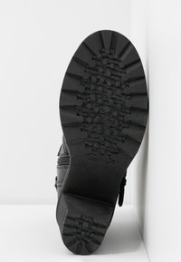 Chaussure noire avec une semelle en caoutchouc texturée comportant des crampons profonds, des matériaux composites et un design plat. Fermeture éclair latérale et détails de boucle visibles.