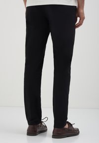 Pantalon noir ajusté, avec une texture lisse et des détails de couture subtils. Assorti à des chaussures en cuir marron à lacets.