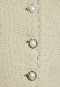 Cremefarbener Stoff mit strukturiertem Gewebe, versehen mit drei großen Knopfverschlüssen mit Perlenmitten und dekorativen silbernen Akzenten.