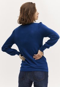 Modra pletena jopica z dolgimi rokavi, rebrastimi manšetami, okroglim izrezom in rahlo napihnjenimi rameni, nosena z temnimi jeans hlačami.