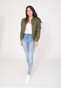 Oliwkowa kurtka puchowa z prążkowanymi mankietami i kołnierzem, zapinana na zamek z przodu, z bocznymi kieszeniami. Połączona z jasnoniebieskimi obcisłymi dżinsami i białymi trampkami.