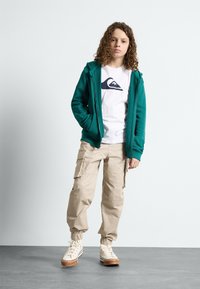 Felpa verde con zip sopra una maglietta bianca con un logo, pantaloni cargo beige con polsini elasticizzati e sneakers beige con suole marroni.