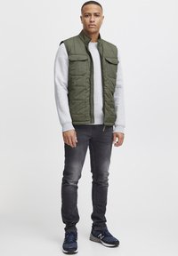 Olivgrüne Steppweste mit hohem Kragen, zwei Vorderaschen, getragen über einem grauen Sweatshirt, kombiniert mit schmal geschnittenen grauen Jeans und blauen Sneakern.