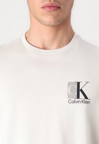 Světle béžové bavlněné tričko s malým černým logem "CK" a textem "Calvin Klein" na levé straně hrudi, kulatý výstřih.