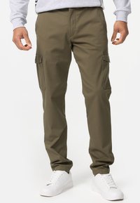Pantalons cargo vert olive avec deux poches latérales, fabriqués en tissu léger ; coupe détendue avec un design à jambes droites, assortis à des baskets blanches.