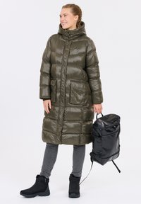 Puffjacka i olivgrön, knälång med en glänsande yta, två framfickor, hög krage, i kombination med svart ryggsäck och stövlar.