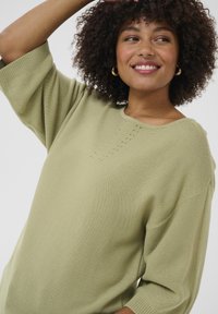 Hellgrüner Strickpullover mit lockerer Passform, weiten Ärmeln und rundem Ausschnitt. Verfügt über eine Reihe kleiner Lochdetails entlang der Brust.