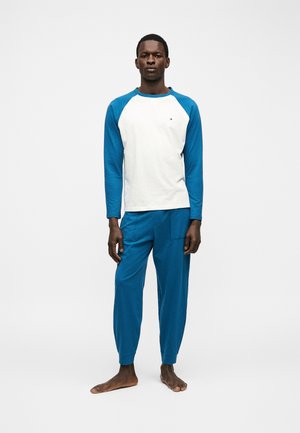 Mężczyzna stoi boso, ubrany w niebieską koszulę raglanową z długim rękawem z białym tułowiem i dopasowane niebieskie spodnie na jednolitym białym tle.