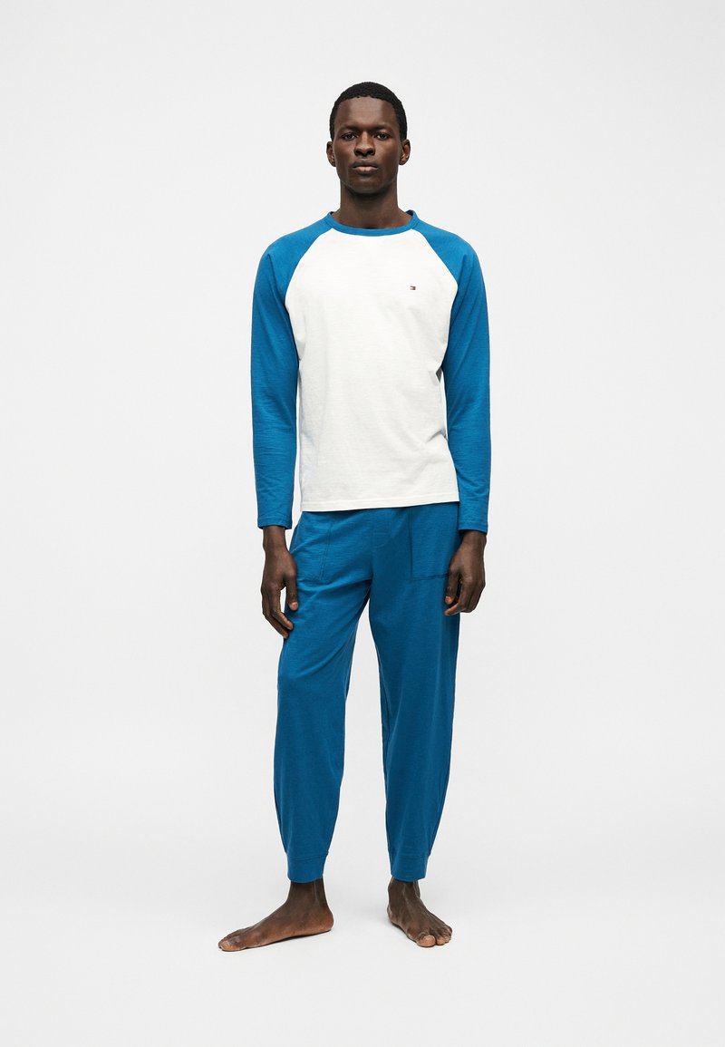 Άντρας που στέκεται ξυπόλητος φορώντας μπλε μακρυμάνικη ραγκλάν μπλούζα με λευκό σώμα και ταιριαστό μπλε παντελόνι σε απλό λευκό φόντο.