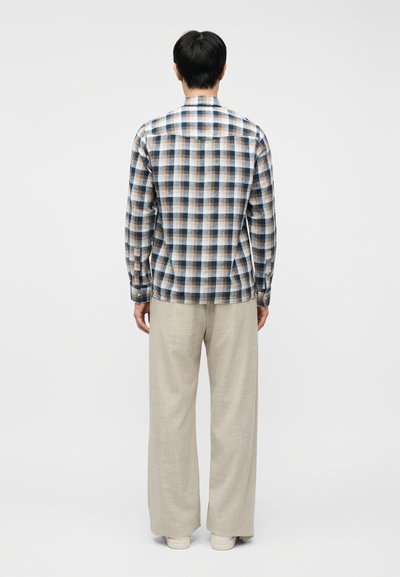 Άνδρας που στέκεται έχοντας την πλάτη του προς τα εμπρός, φοράει μπλε, καφέ και λευκό πουκάμισο με μακριά μανίκια σε καρό σχέδιο, μπεζ παντελόνι φαρδύ και λευκά παπούτσια.