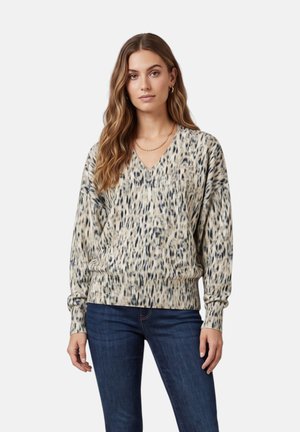 Donna che indossa un maglione beige e nero a V con maniche lunghe e jeans blu, in piedi su uno sfondo bianco uniforme.