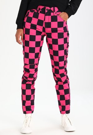 Person iført pink og sort ternede bukser, sort top og hvide høje sneakers, stående med den ene hånd i lommen.