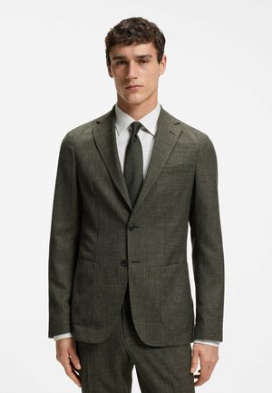 Uomo che indossa un abito verde scuro texturizzato con due bottoni, camicia bianca e cravatta scura, in piedi contro uno sfondo bianco semplice.