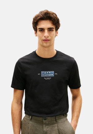 Joven con una camiseta negra de Tommy Hilfiger con el texto del logo y pantalones de color verde oliva, de pie contra un fondo liso y claro.