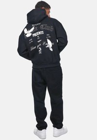 Sort hættetrøje med hvid grafiktekst og fugle på bagsiden. Sammen med sorte bukser og hvide sneakers. Blødt materiale, afslappet pasform.