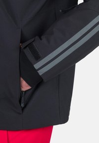 Giacca nera con strisce riflettenti grigie, dotata di una tasca con zip e chiusura a strappo sulla manica per una vestibilità sicura e regolabile.