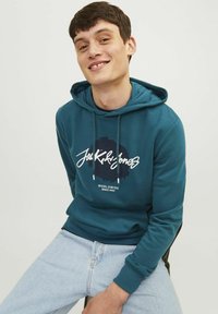 Sudadera con capucha color teal, con un gráfico en la parte frontal y cordones ajustables. Llevada con jeans de un azul claro, que presentan una textura suave y un ajuste relajado.
