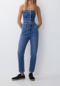 Haut en denim sans bretelles avec boutons sur le devant, associé à un jean bleu taille haute. Baskets noires avec semelles en caoutchouc. Texture lisse du denim sur l'ensemble.