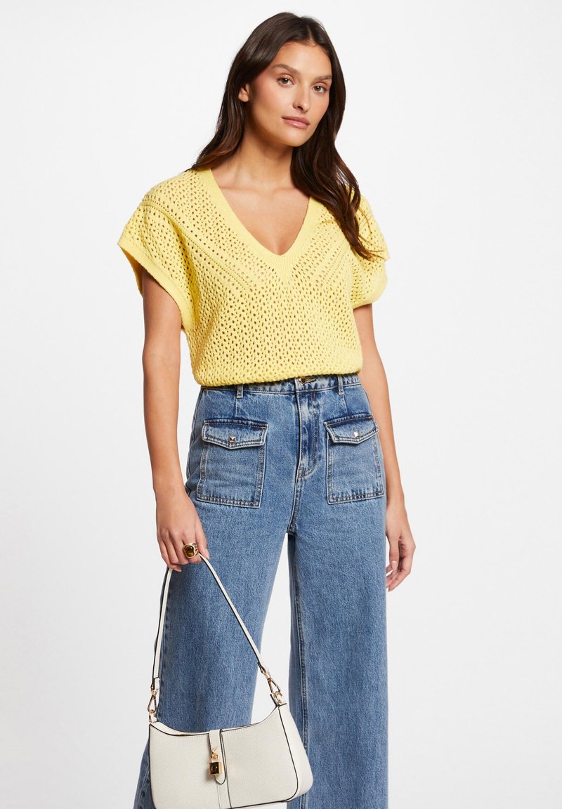 Top giallo lavorato a maglia con scollatura a V e maniche corte abbinato a jeans larghi in denim blu a vita alta e una piccola borsa bianca.