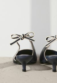 Tummansinisen nahkakenkiä matalalla, kaarevalla korolla, joissa on vaalean beigeä satiinirusettia takana ja sievästi viimeistellyt reuna. Silkkinen pinta, minimalistinen muotoilu.