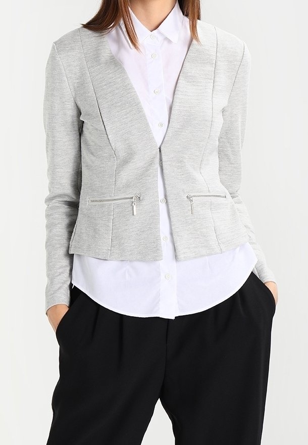 Femme portant un blazer zippé gris clair sur une chemise blanche boutonnée et un pantalon plissé noir, les mains dans les poches.