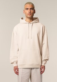 Hoodie beige en tissu doux, avec une poche kangourou, une capuche avec cordon et des poignets côtelés. Porté par-dessus un pantalon clair.
