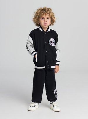 Giacca varsity nera con maniche grigie, caratterizzata da una toppa con una scimmia e accenti a strisce bianche. Abbinata a pantaloni della tuta neri e sneakers bianche.