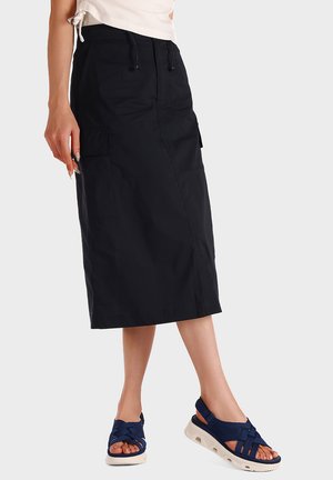 Donna che indossa una gonna midi nera con tasche laterali, un top bianco annodato e sandali aperti blu navy con suola bianca su sfondo bianco.