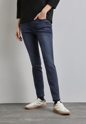 Jeans en denim bleu foncé au design ajusté, avec une taille mi-haute et des coutures discrètes, associés à des baskets blanches et beige.