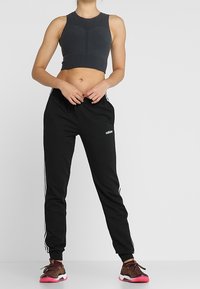 Černé sportovní kalhoty s bílými bočními pruhy a logem Adidas, kombinované s tmavě šedým sportovním podprsenkou, která zdůrazňuje p�řiléhavý design.