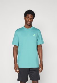 Camiseta de manga corta en color turquesa, hecha de una tela suave, con un pequeño logo de tres rayas en verde claro en el lado izquierdo del pecho. Pantalones cortos negros.