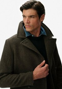 Abrigo marrón de espiga con un gran cuello, llevado sobre un suéter negro y una camisa de mezclilla. Texturas notables y ajuste a medida.