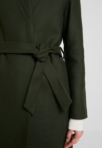 Cappotto verde oliva con un tessuto testurizzato, design avvolgente e una cintura legata in vita. I polsini sono visibili in un colore più chiaro.