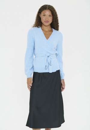 Vrouw met krullend haar, gekleed in een lichtblauwe wikkeltrui die op de taille is vastgebonden en een lange zwarte rok, staande tegen een witte achtergrond.