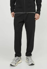 Black jogger pants made of soft fabric, featuring a tapered fit, side seam pockets, and contrasting stitching. Paired with gray athletic shoes.