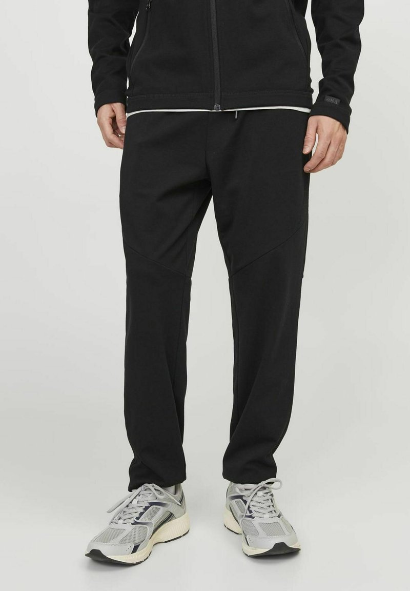 Black jogger pants made of soft fabric, featuring a tapered fit, side seam pockets, and contrasting stitching. Paired with gray athletic shoes.