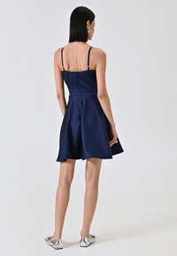 Robe en satin bleu marine avec des bretelles fines, jupe évasée et fermeture éclair au dos. Associée à des chaussures plates en métal argenté. Texture lisse.
